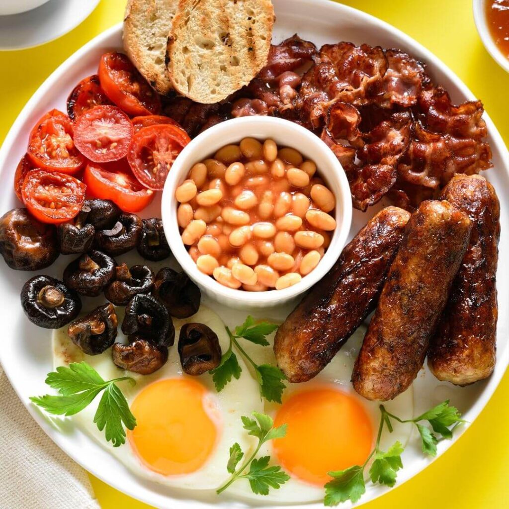 A full English breakfast plate filled with two fried eggs, grilled baby mushrooms, grilled cherry tomatoes, toast, crispy bacon and three sausages with a white dish filled with baked beans in the center of the plate.