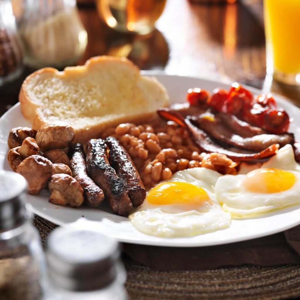 A full English breakfast with fried eggs, sausage, back bacon, baked beans, tomatoes, grilled mushrooms and toast sitting on a white plate