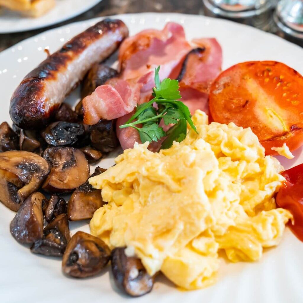 A white plate holding a full English breakfast of scrambled eggs, grilled tomato, back bacon, sausage and mushrooms.