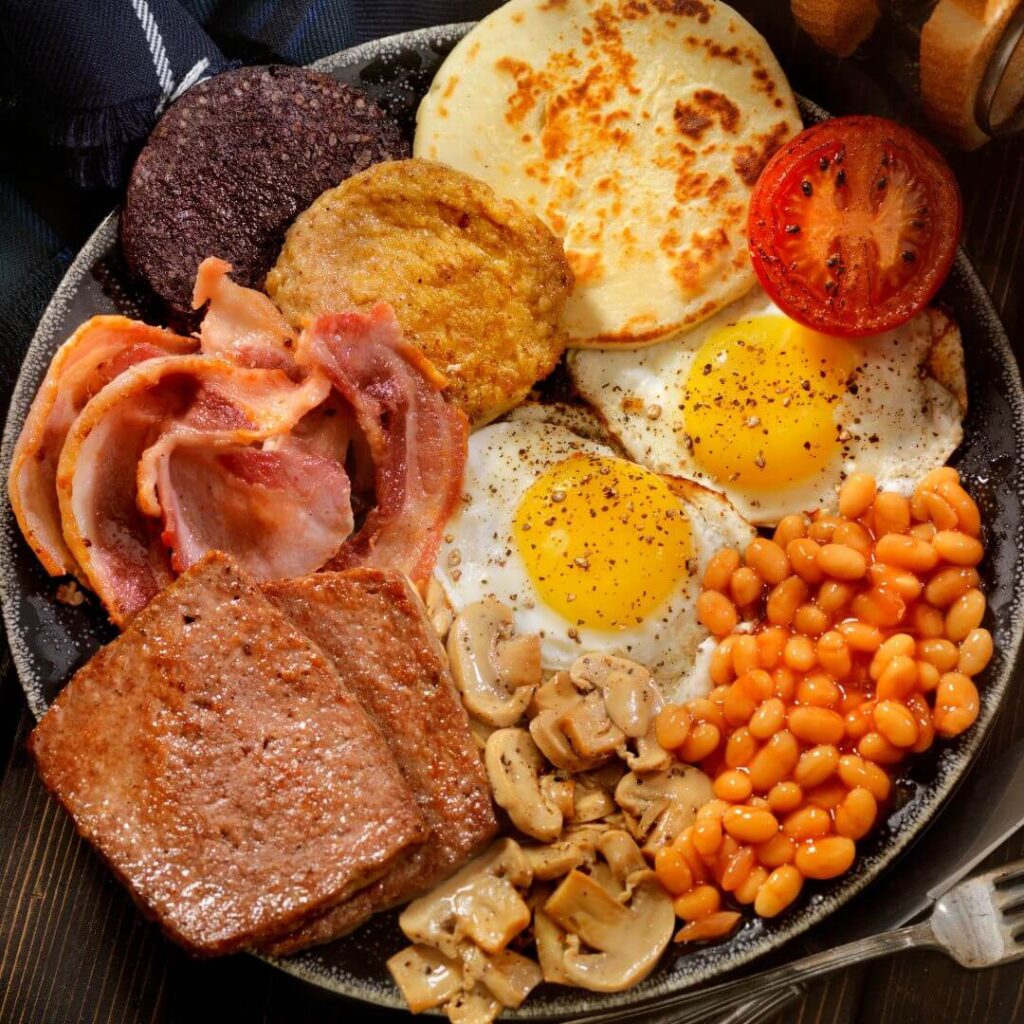 A traditional breakfast in Scotland. A black plate filled with food including Lorne sausage, back bacon, black pudding, white pudding, tattie scones, grilled tomato, fried eggs, baked beans and mushrooms.