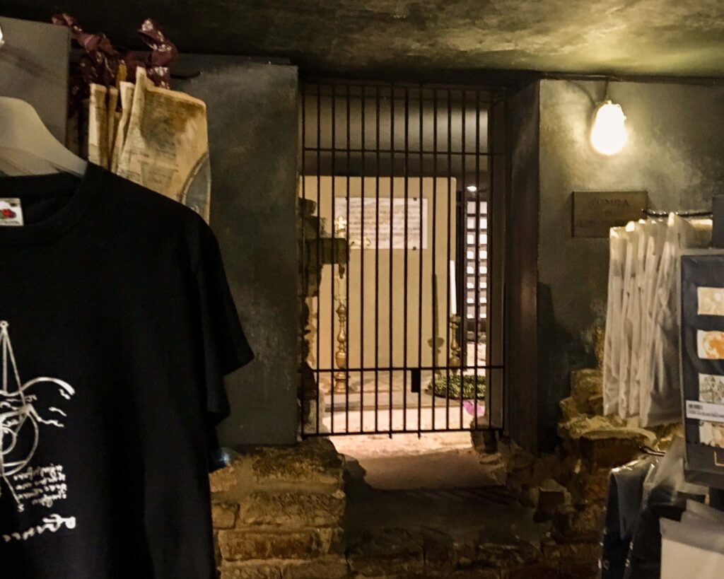 The crypt under the Florence Cathedral where Filippo Brunelleschi is buried. The crypt has a simple metal gate, and you can see candlesticks and a wreath behind. In front of the gate are racks of gift shop merchandise.