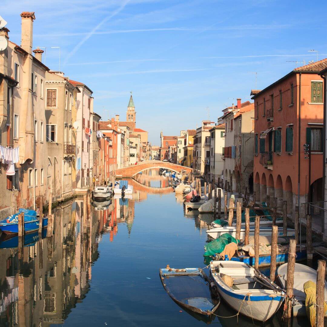 The main canal through Chioggia Italy, with a bridge crossing over and boats along the sides. A belltower stands tall in the background.