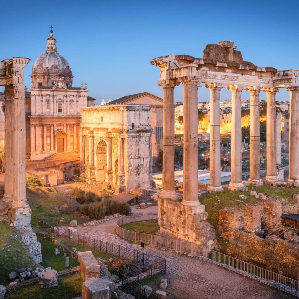 An evening view of the Roman Forum and Arch of Titus. The structures are illuminated, making them stand out against the darkening sky.