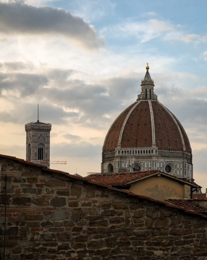 The dome of the Florence Cathedral can be seen over the rooftops of Florence at sunset, with a blue sky starting to turn orange and pink behind