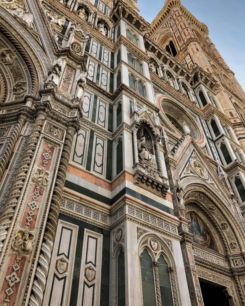 A detailed view of the facade of the Florence cathedral. The pink, green and white marble is highly detailed, with geometric designs as well as multiple statues and detailing to give the cathedral visual interest
