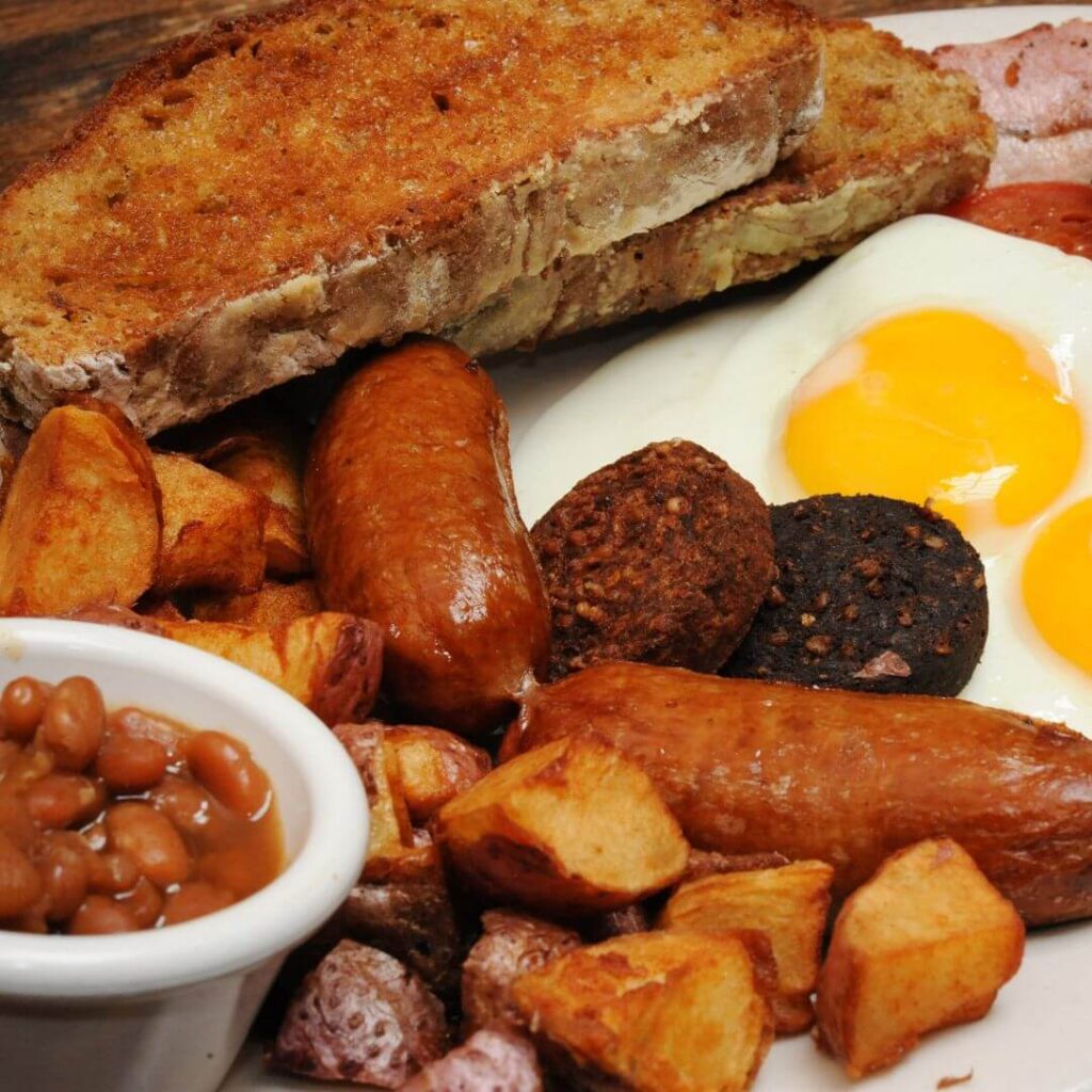 Close up of a traditional Irish breakfast including fried eggs, irish rashers, Irish sausage, black pudding, white pudding, potatoes, irish soda bread and a small white dish of baked beans.