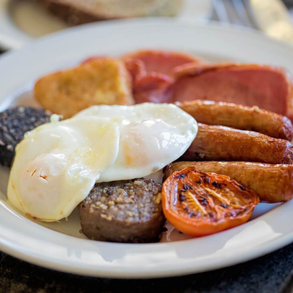 Full Irish breakfast on a white plate, including two fried eggs, two Irish rashers, three Irish sausages, one grilled tomato, black pudding, white pudding, and a hash brown.