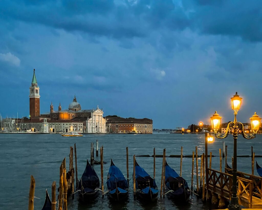 The church of San Giorgio Maggiore at dusk. Gondolas can be seen at the edge of the Piazza San Marco, with the church across the canal. A lit Venetian streetlight adds to the mood.