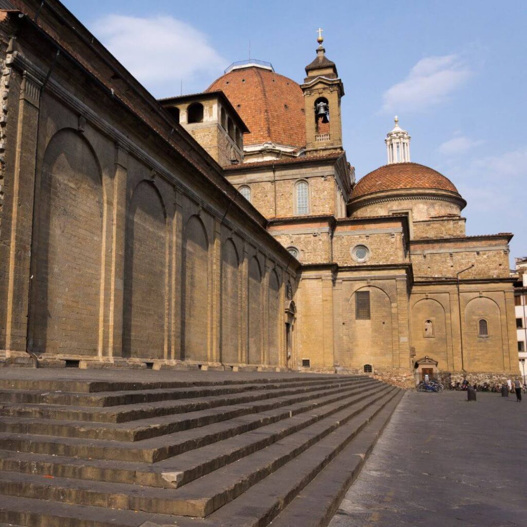 The exterior of San Lorenzo in Florence, Italy. Steps lead up to the front and side of the church, with the domes of the Medici Chapels in the background.