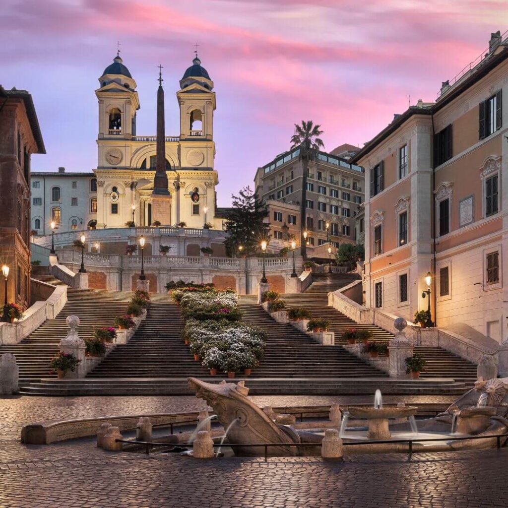 The Spanish Steps at sunset in Rome Italy. The clouds in the sky are turning pink and purple, and all the streetlights are on, illuminating the steps.