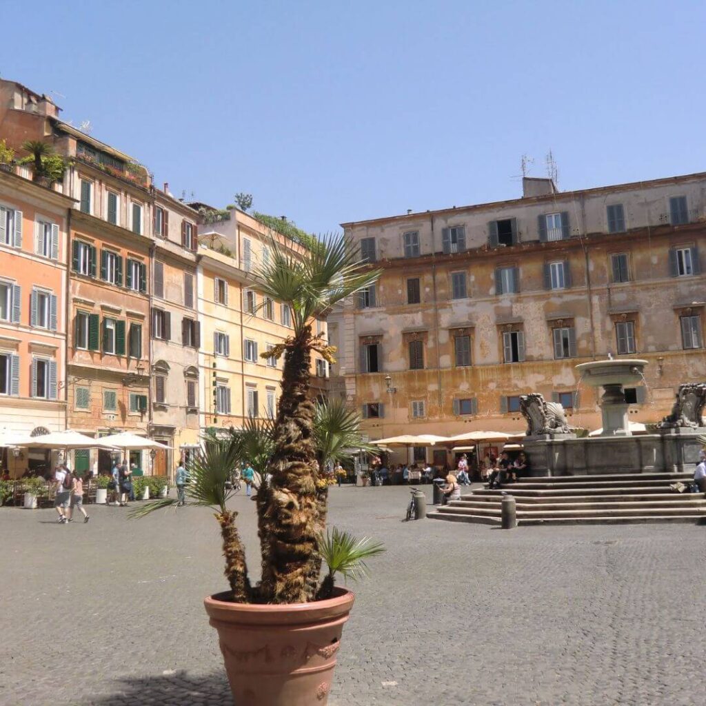 A piazza in the Trastevere area of Rome, with a fountain in the middle and restaurants with tables and umbrellas outside around the edge.