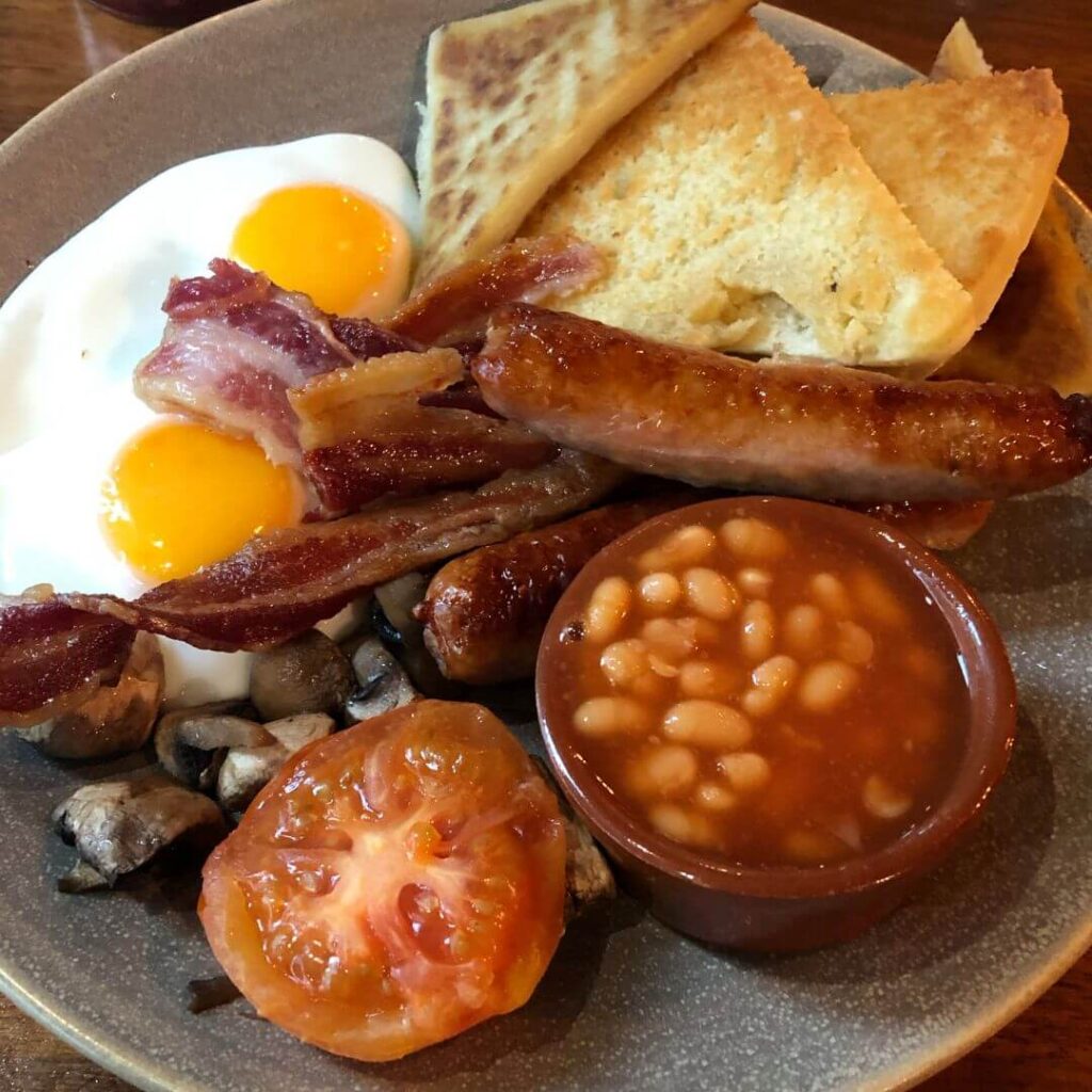 Close up image of an Ulster Fry on a grey plate with soda farls, boxty, two fried eggs, two Irish rashers, two Irish sausages, one grilled tomato, mushrooms and a small brown dish of baked beans. An Ulster Fry is what a traditional Irish breakfast is called in Northern Ireland.