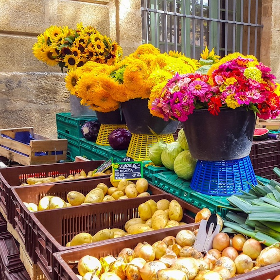 A table full of produce bins of onions and potatoes with colorful flower bouquets in buckets on a shelf above