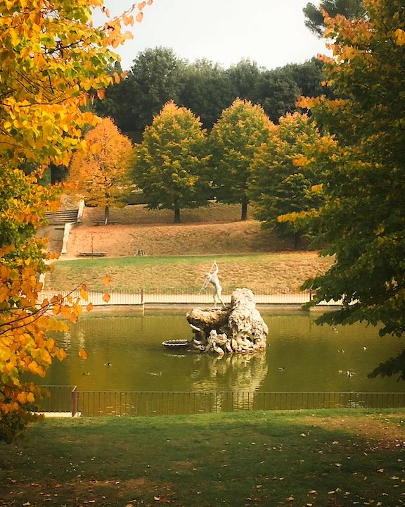 A statue surrounded by green grass with the leaves just beginning to turn orange and yellow in Fall at the Boboli Gardens