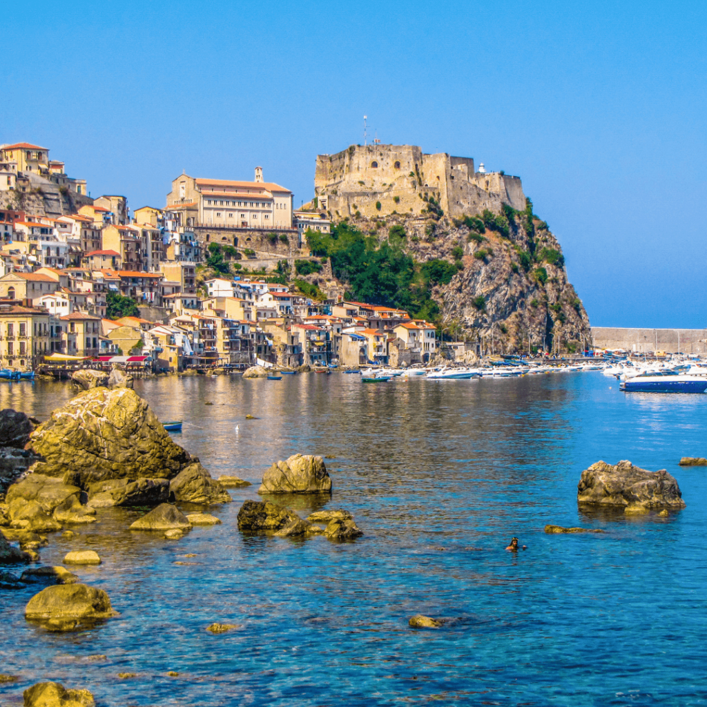 A view of Scilla from the water with the town coming down the hill to the ocean's edge and large rocks in the foreground