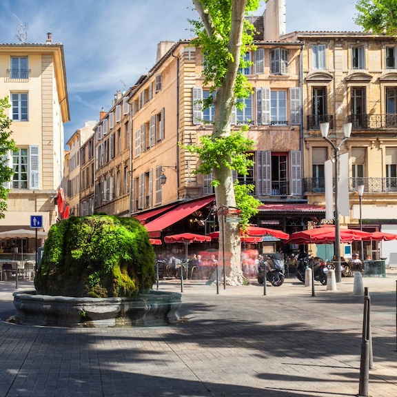 A view of the Cours Mirabeau with red umbrellas over sidewalk tables and green trees along the street.