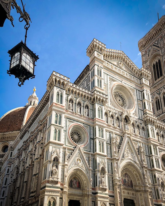 The intricate facade of the Florence cathedral, or Duomo against a blue sky with a hanging lantern in the foreground