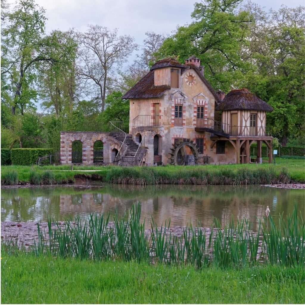 The exterior of the Queens Hamlet at Versailles on a cloudy day, reflected in a still pond