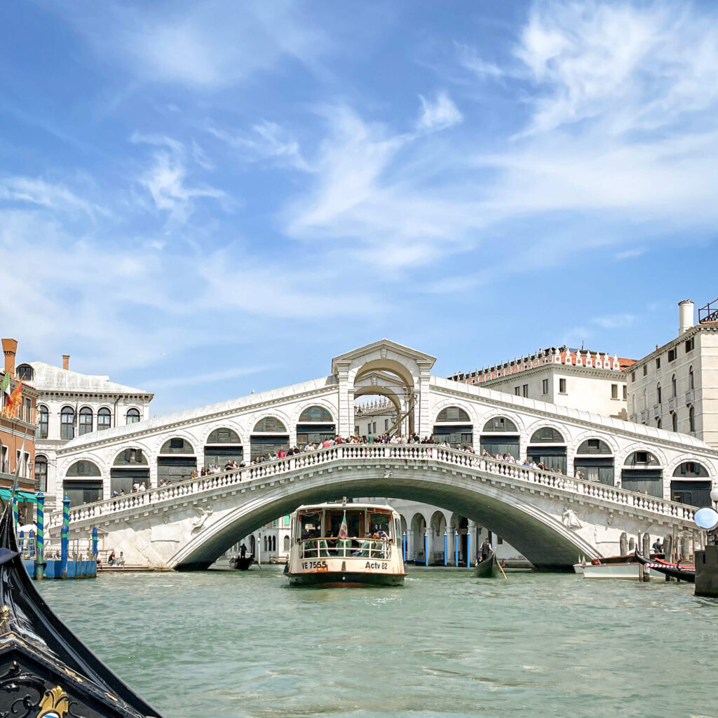 View of the white arches and shops of the Rialto with a boat sailing under the bridge as viewed from a gondola