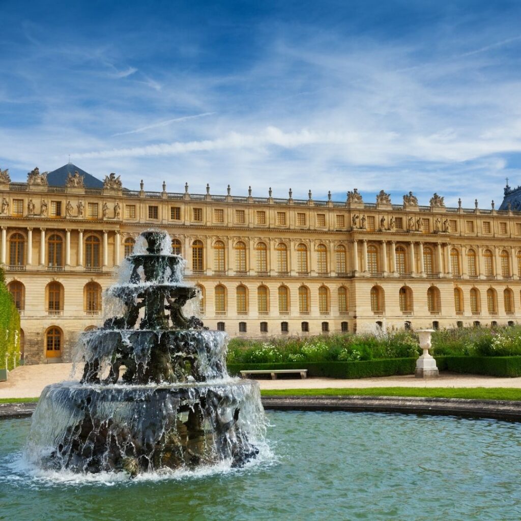 An external view of Versailles against a blue cloudy sky with wispy white clouds and a running fountain in the foreground