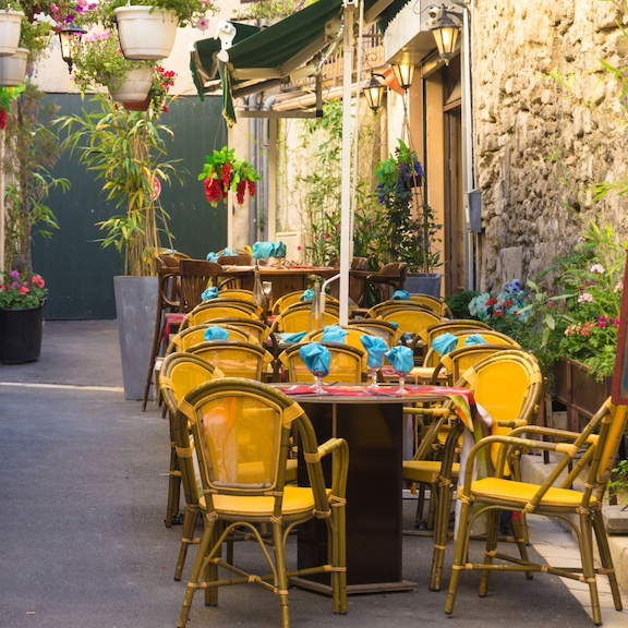 A sidewalk cafe in Old Aix with yellow chairs at a round table with red placemats and water goblets with folded turquoise napkins inside, one of the most common things to do in Aix-en-Provence