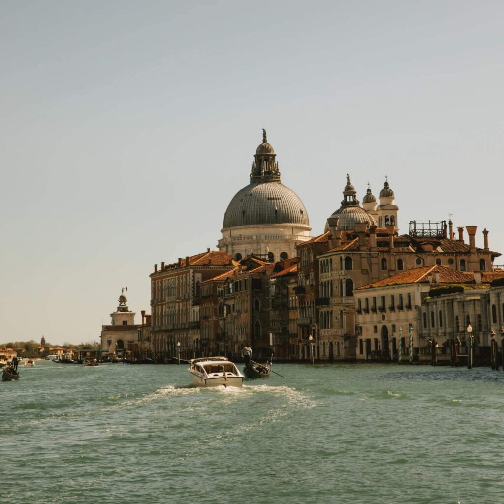 View across the Venetian Grand Canal of the tan and brown buildings with red roofs and the dome of a church silhouetted against a dull blue sky