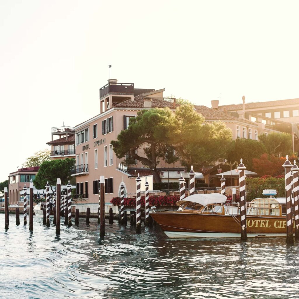 Exterior view of the canal-side entrance to Hotel Cipriani with its pinky-beige walls rising above the green tree tops with the Hotel Cipriani shuttle boat next to red and white striped pilings in the foreground.