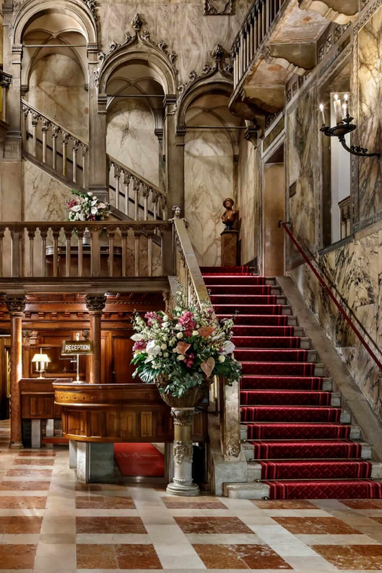 Ornate marble-floored interior of the reception area at Hotel Danieli with a red-carpeted staircase to the second floor and the reception desk tucked next to the stairs.