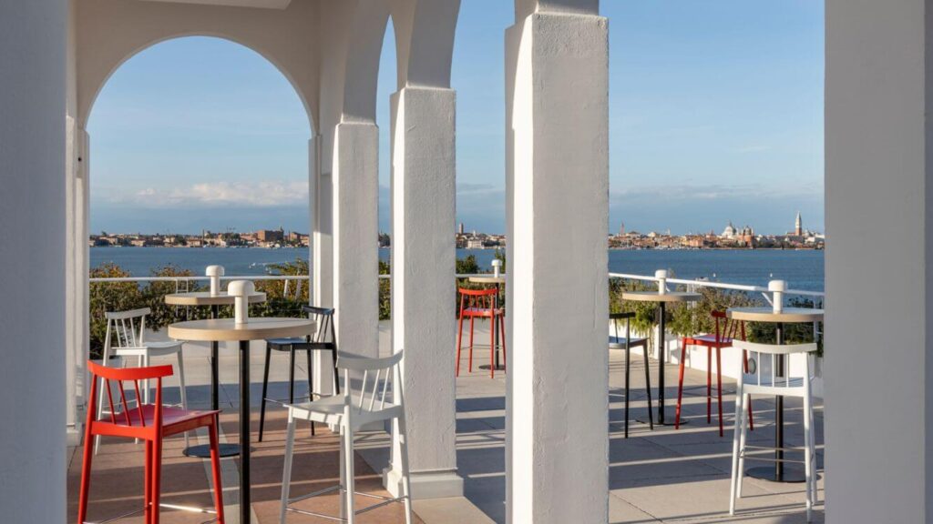 White columns and archways over the Terrace filled with high tables and brightly colored chairs overlooking the Venetian lagoon towards the main part of Venice