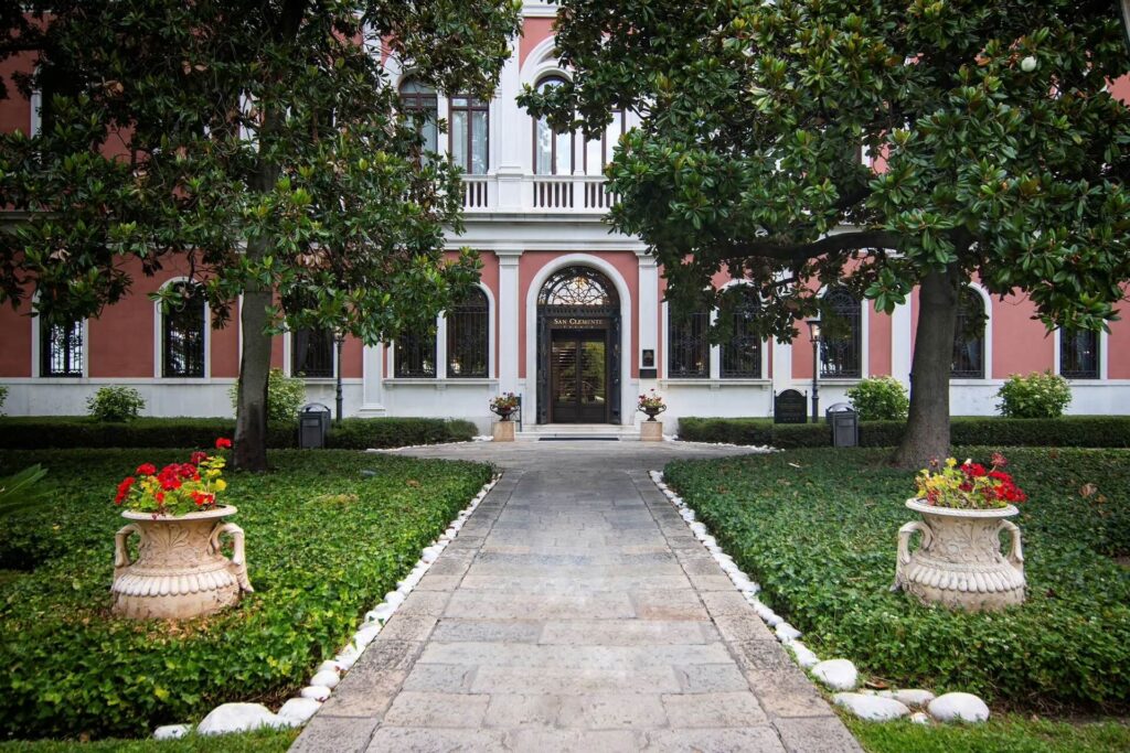 Tan plant-filled urns and green leafed trees flanking the grey stone walkway leading to the entrance of San Clemente Palace